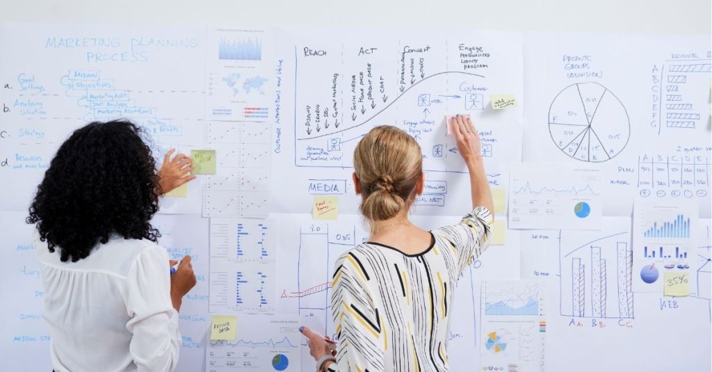 Two women in front of a bulletin board filled with graphs and charts
