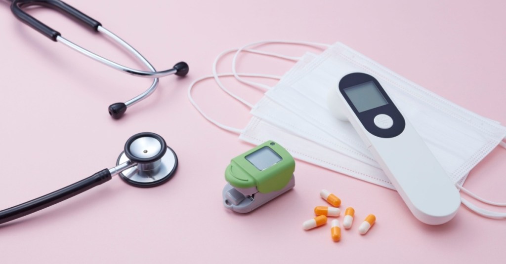 A stethoscope, oral thermometer, finger oximeter, pills, and face masks lying down in front of a pink backdrop