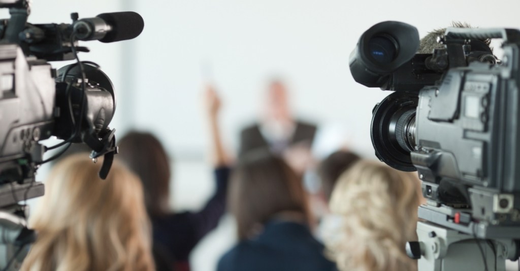 Two broadcast cameras in front of a blurred image of a person raising their hand