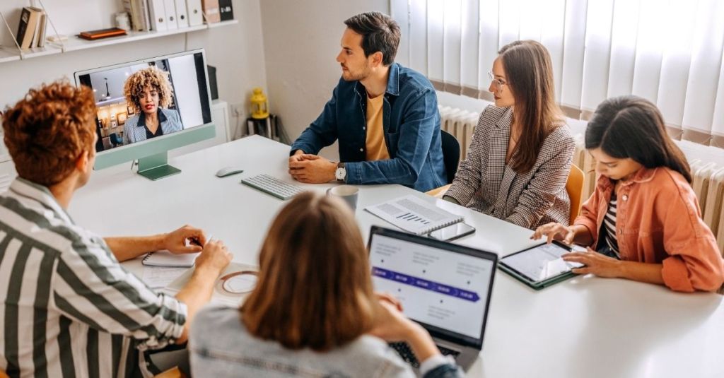 Team members sitting at a table looking at their manager who's speaking from a computer