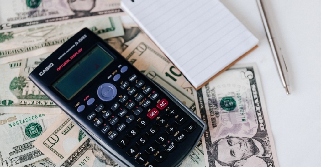 A calculator on top of a pile of money with a notepad beside it