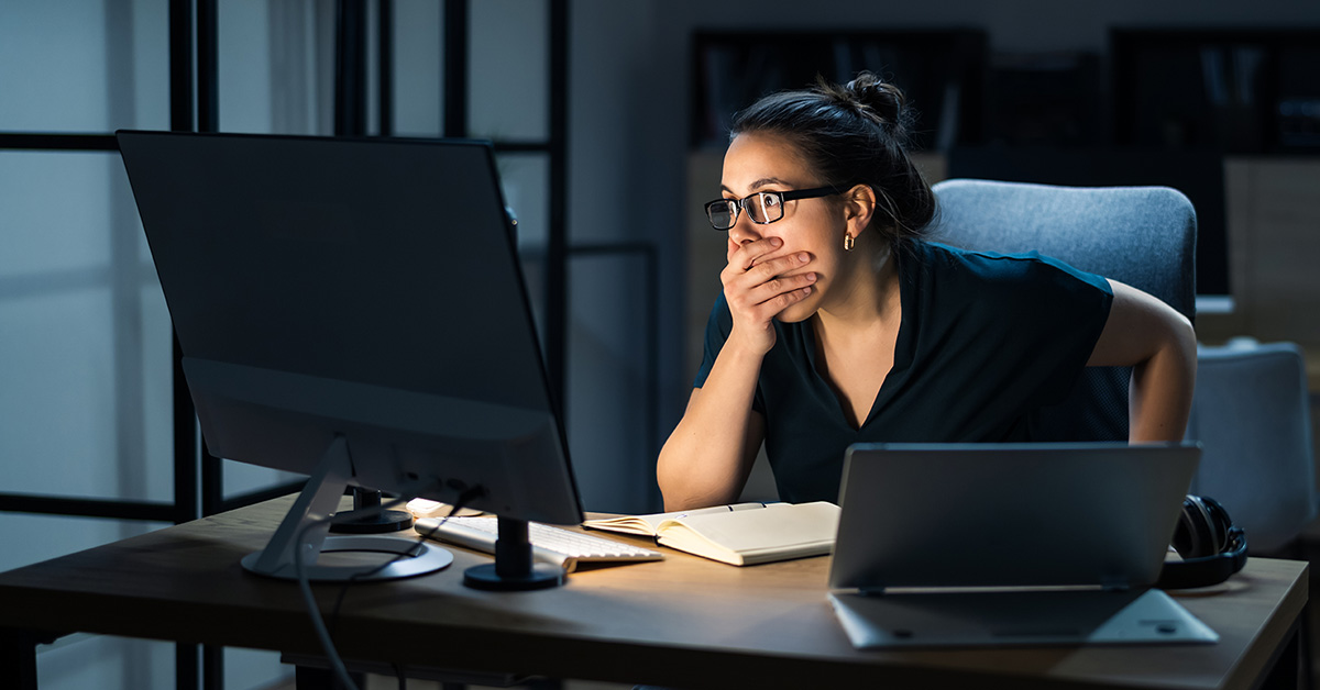 A woman looking shocked with her hand over her mouth, looking at a computer in a dark room