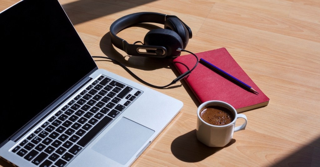 A laptop, headphones, a cup of coffee, and a notebook lying on a table.