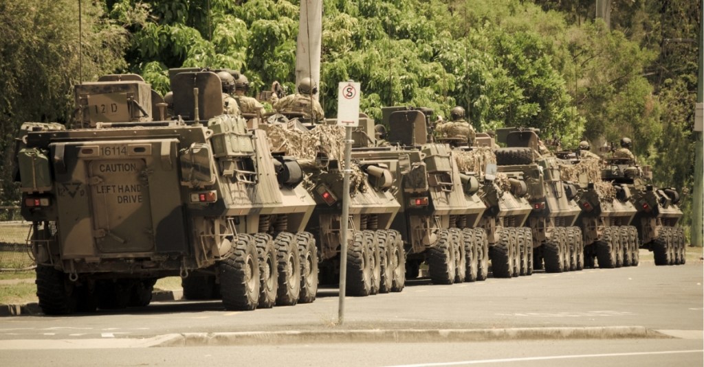 Seven military army tanks driving down a street with people inside.